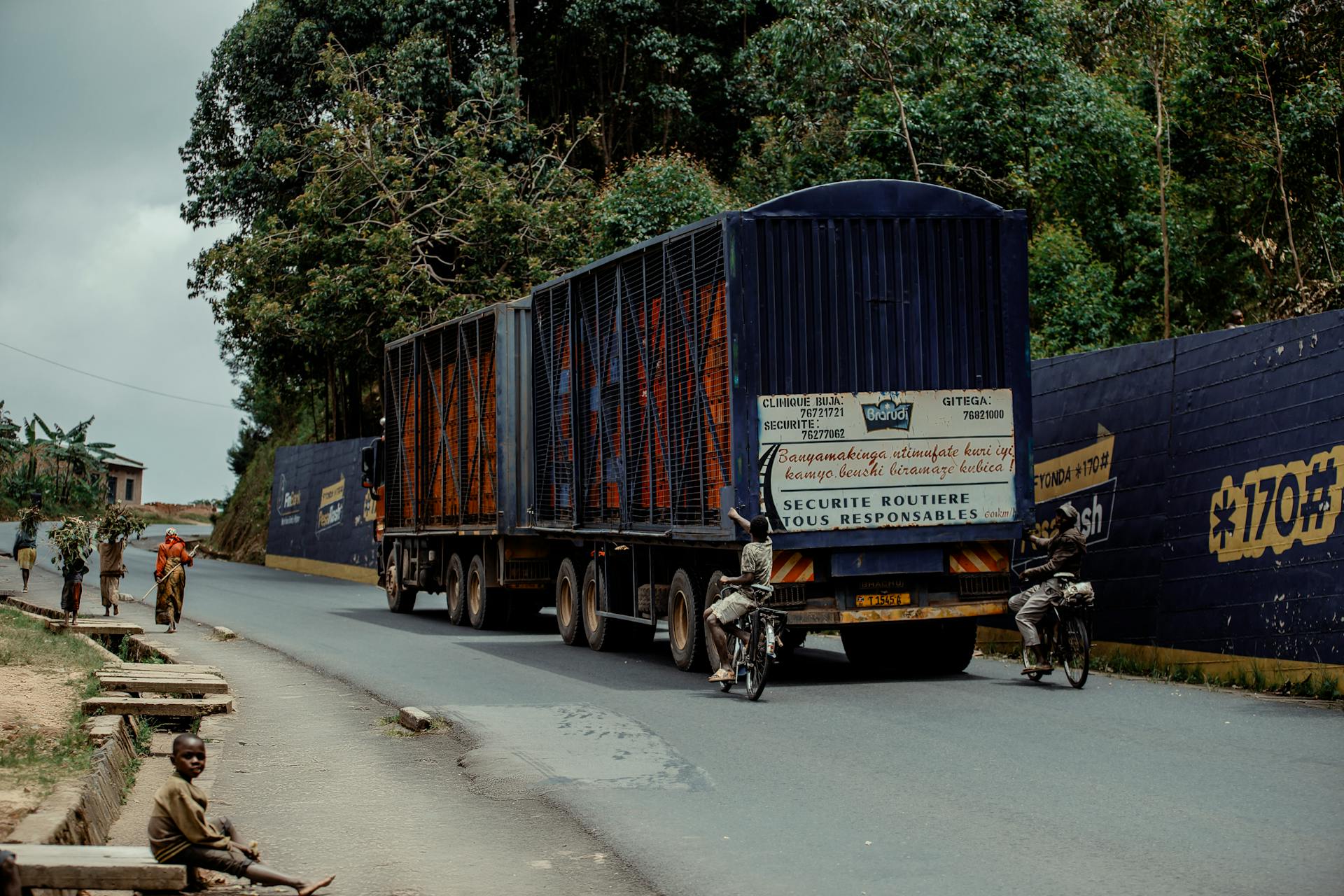 A large truck travels a rural road with cyclists and pedestrians nearby, featuring lush greenery.