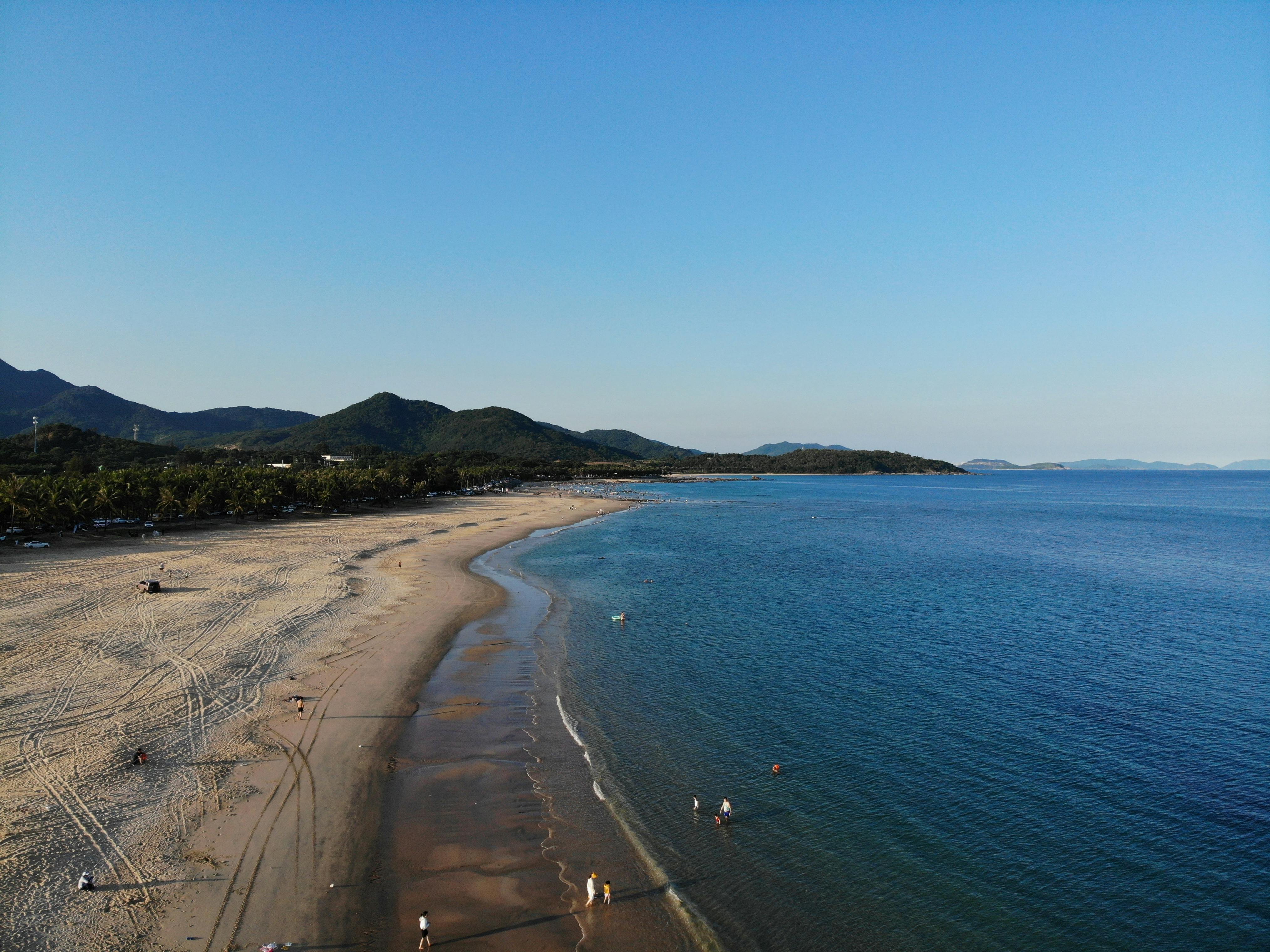 A stunning aerial view of a tranquil beach with gentle waves and distant mountains under a blue sky.