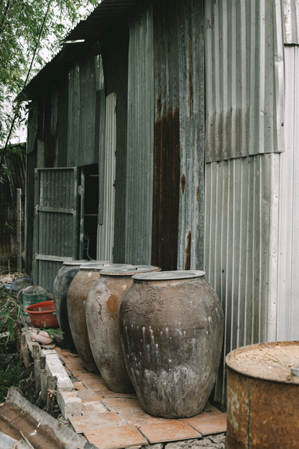 Bảng Giá Hạt Macca Mới Nhất Hiện Nay - Mua Ở Đâu Giá Tốt? 4 Three large rustic jars lined up beside an old corrugated metal shed, reflecting rural Vietnamese architecture.