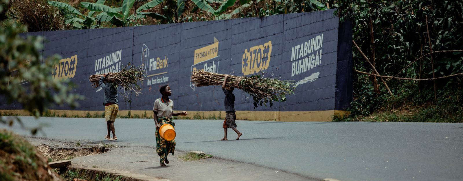 Three individuals walking along a road carrying firewood atop their heads.