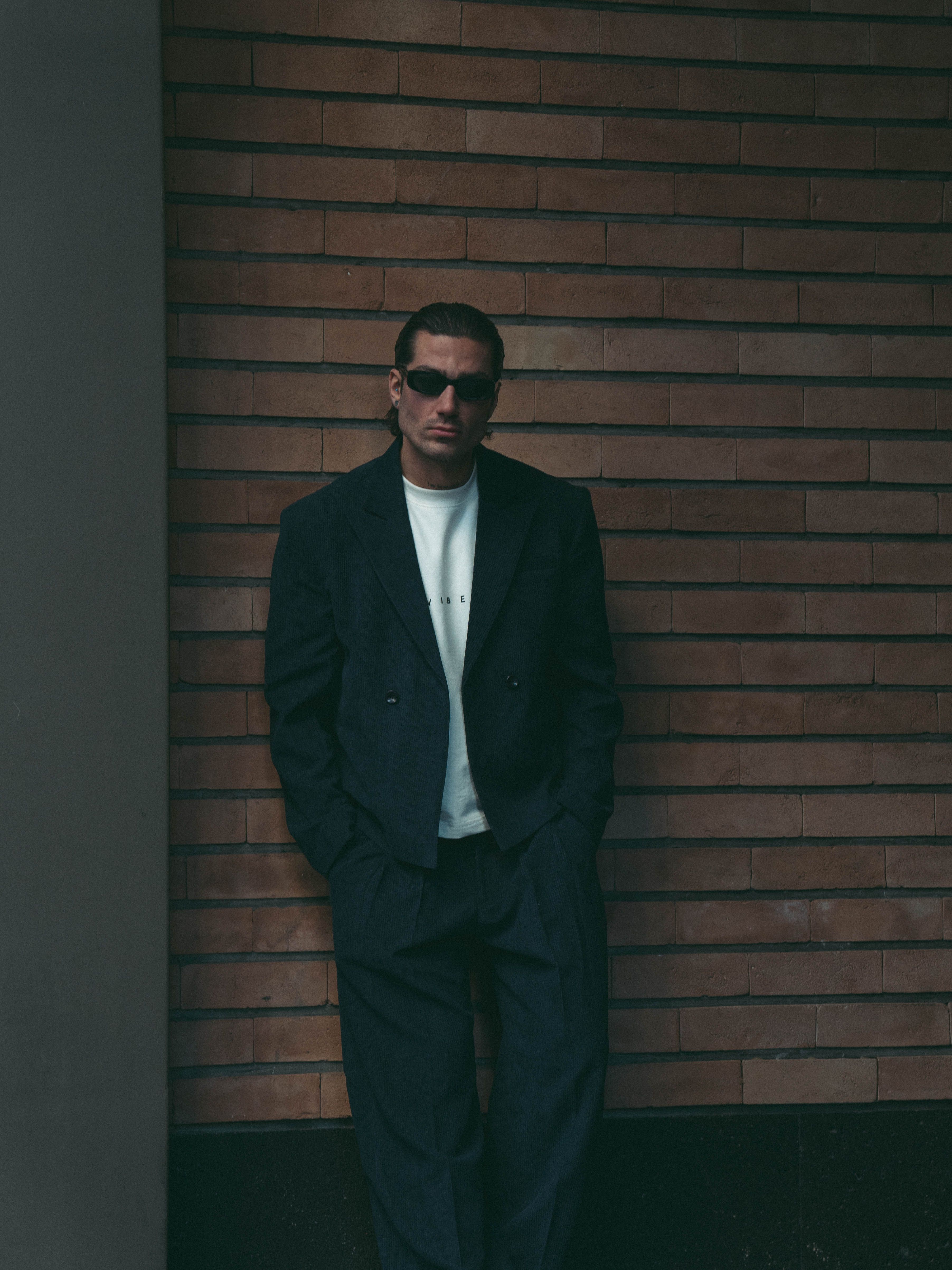 Man wearing sunglasses and a suit stands confidently against a brick wall.