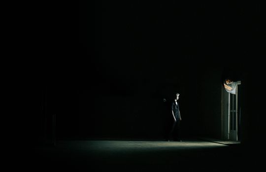 A lone person stands in dim light near a door in a dark room, highlighting isolation.
