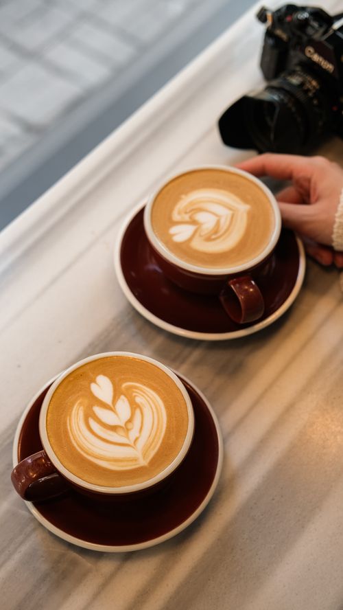 Free Two latte art coffees on a marble table with a hand and camera nearby, perfect for cafe scenes. Stock Photo
