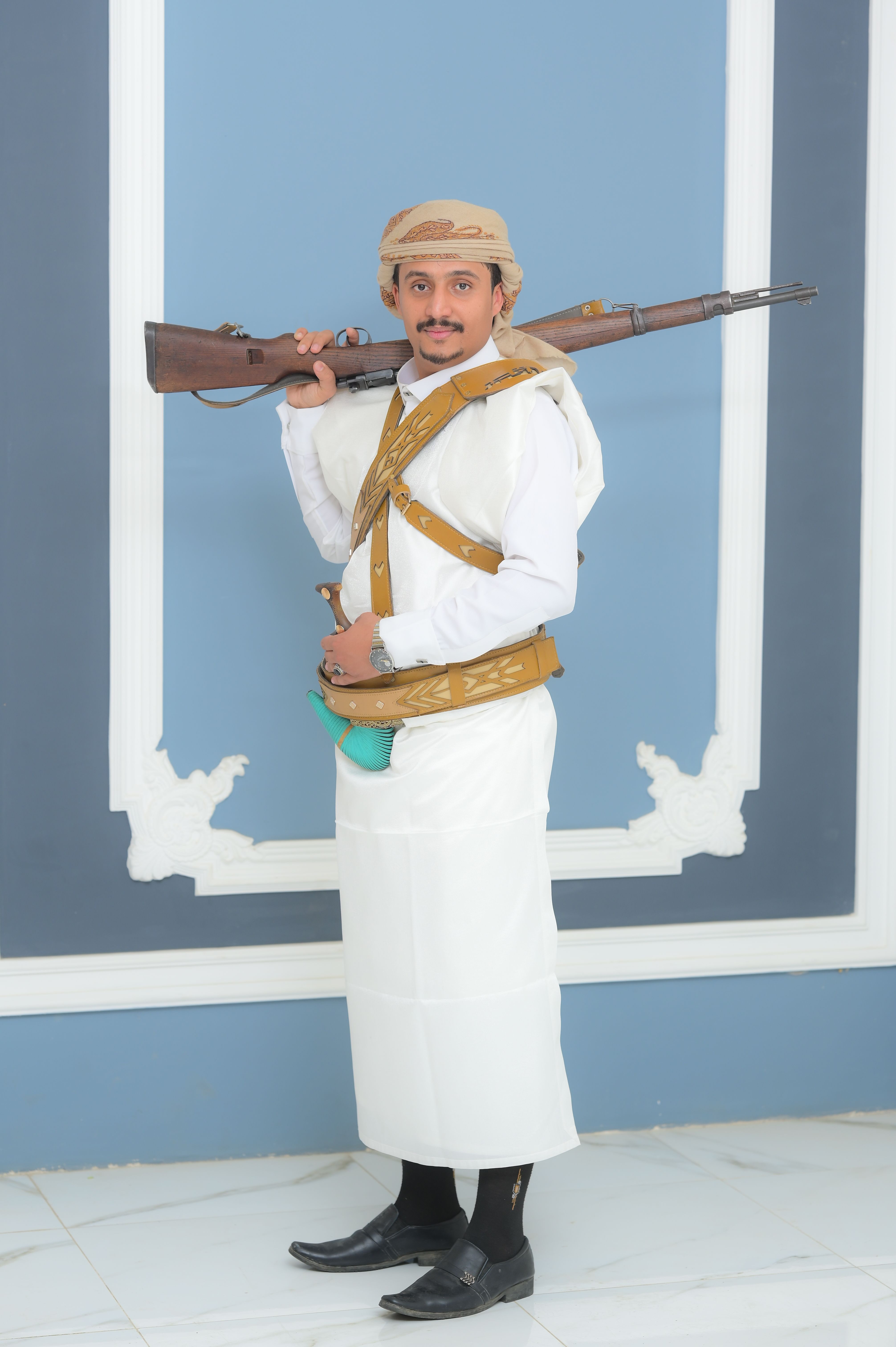 A Yemeni man in traditional attire holding a rifle, showcasing cultural heritage indoors.