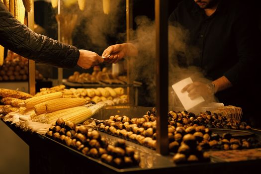 Street food vendor exchanging money at night market with corn and chestnuts under warm light.