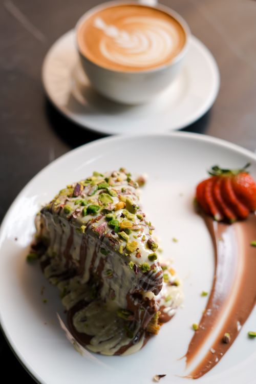 Free Tempting chocolate cake slice topped with pistachios beside latte art coffee on table. Stock Photo