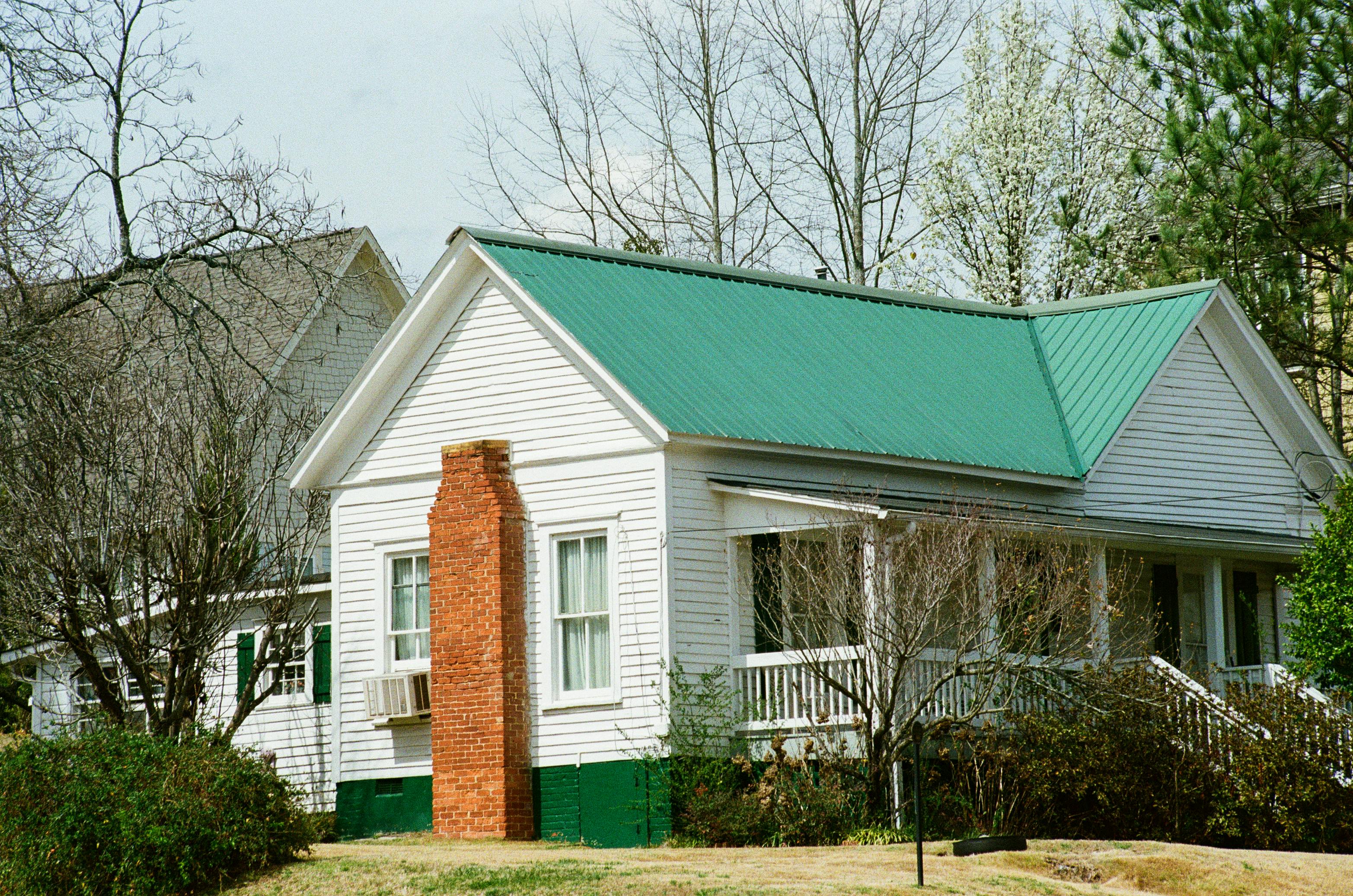 House with green metal roof and brick chimney amidst spring foliage