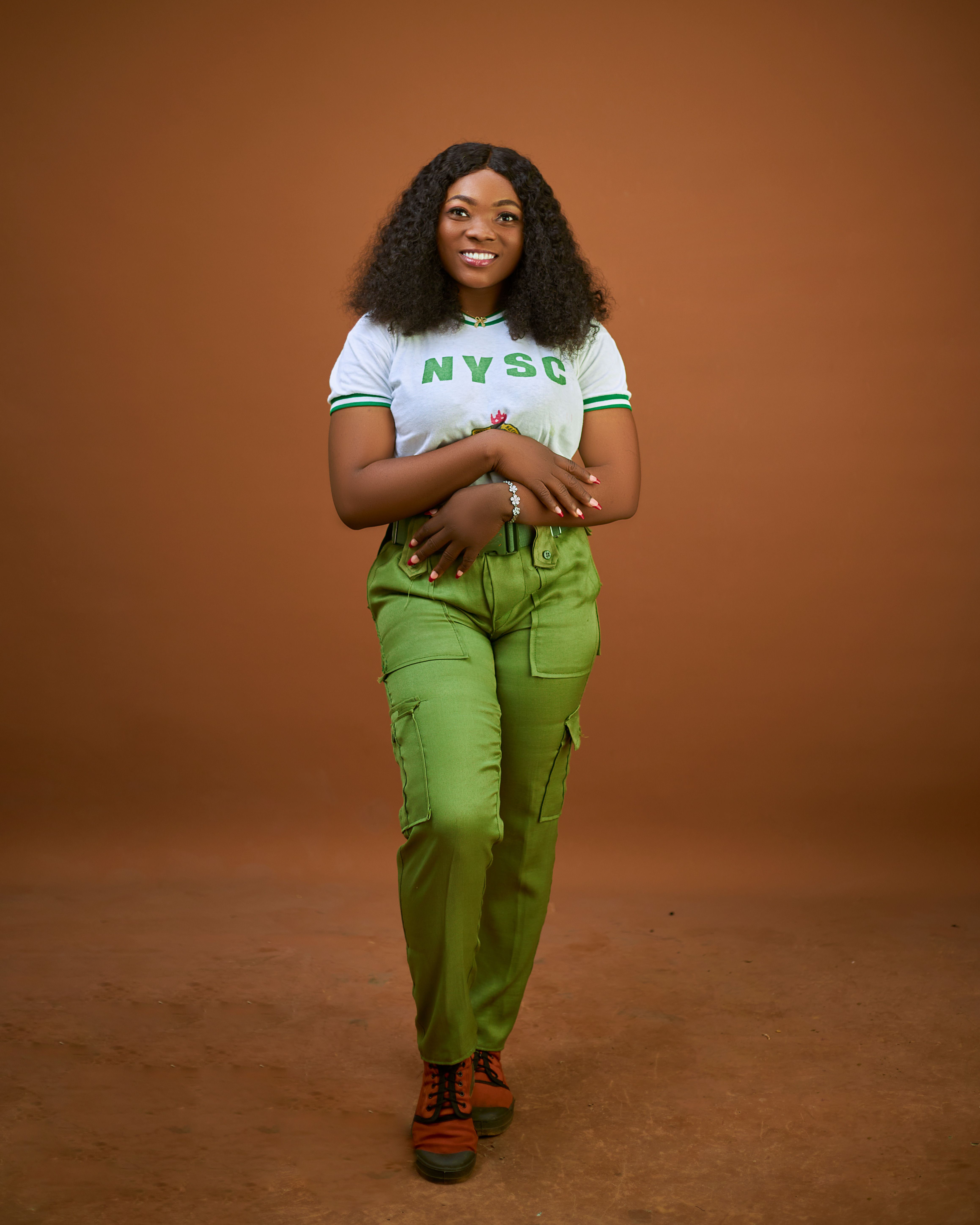 Smiling young woman in NYSC uniform poses confidently against a warm brown backdrop.