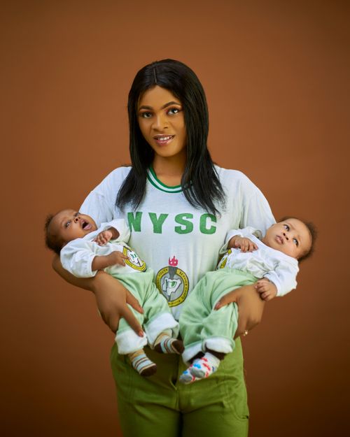 Free A smiling woman in NYSC uniform holding twin babies against a brown backdrop. Stock Photo