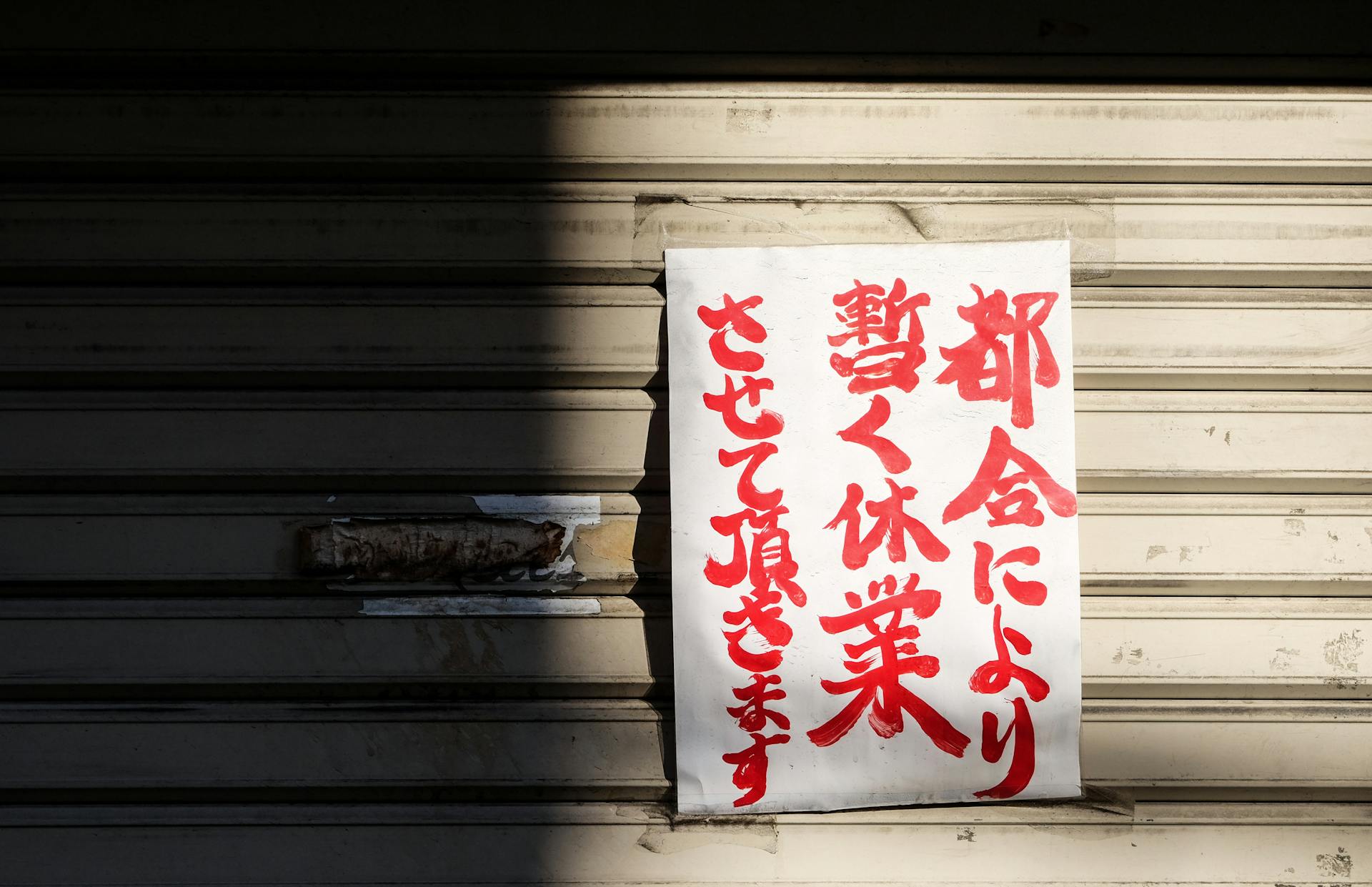 A handwritten Japanese closure sign on a metal storefront shutter, partially shadowed.