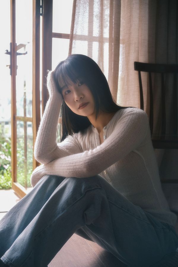 A young woman in casual attire sits pensively near a sunlit window inside a home.