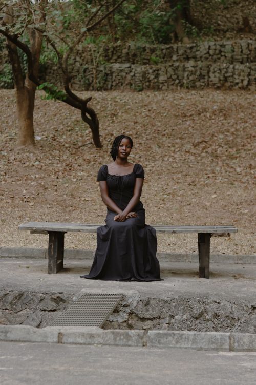 Free A woman in a black dress sitting on a bench in a serene outdoor setting. Stock Photo