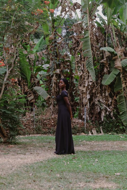 Free A woman in a long black dress stands gracefully in a lush tropical garden setting, surrounded by greenery. Stock Photo