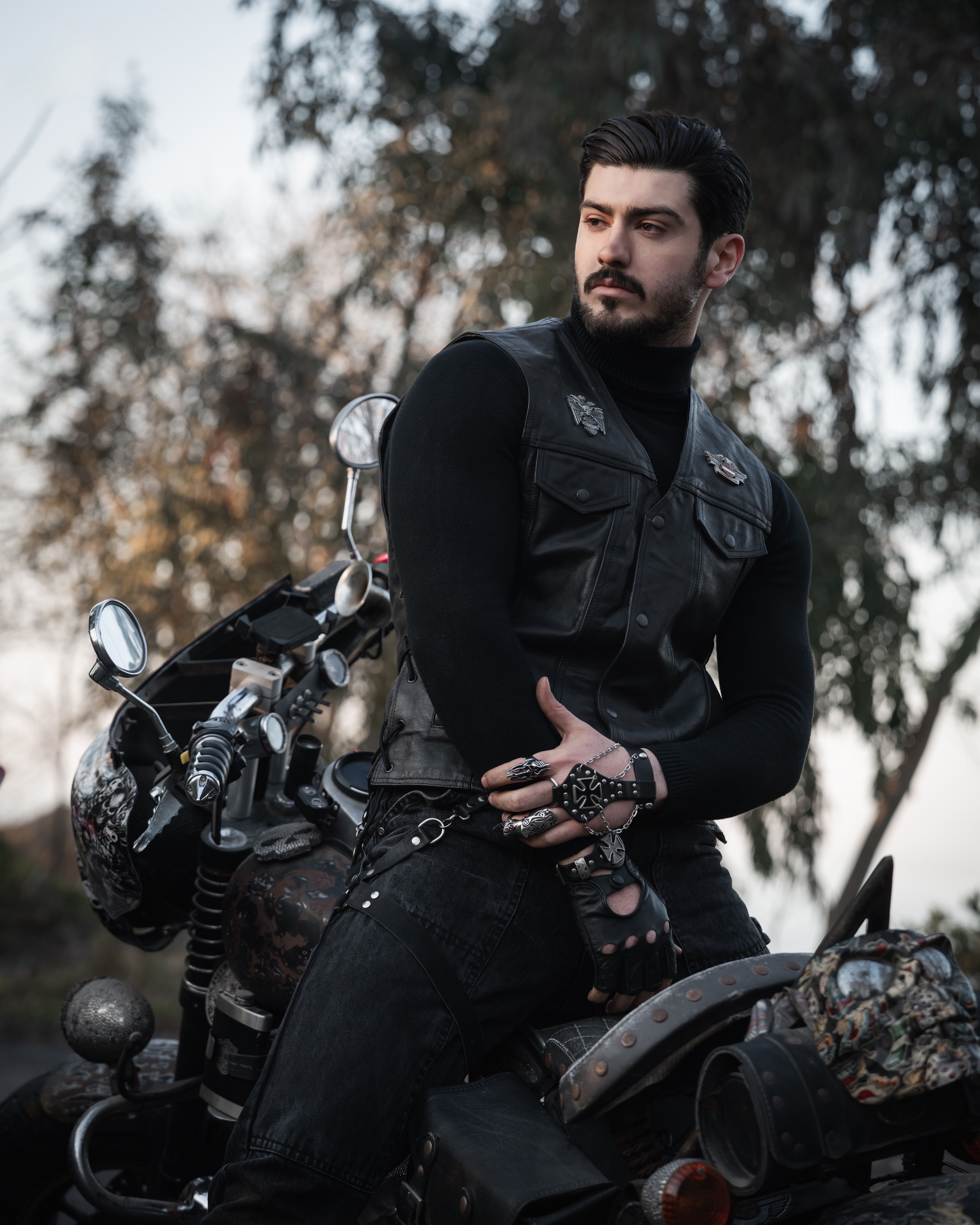 Man in black leather vest posing confidently on a motorcycle in a natural setting.