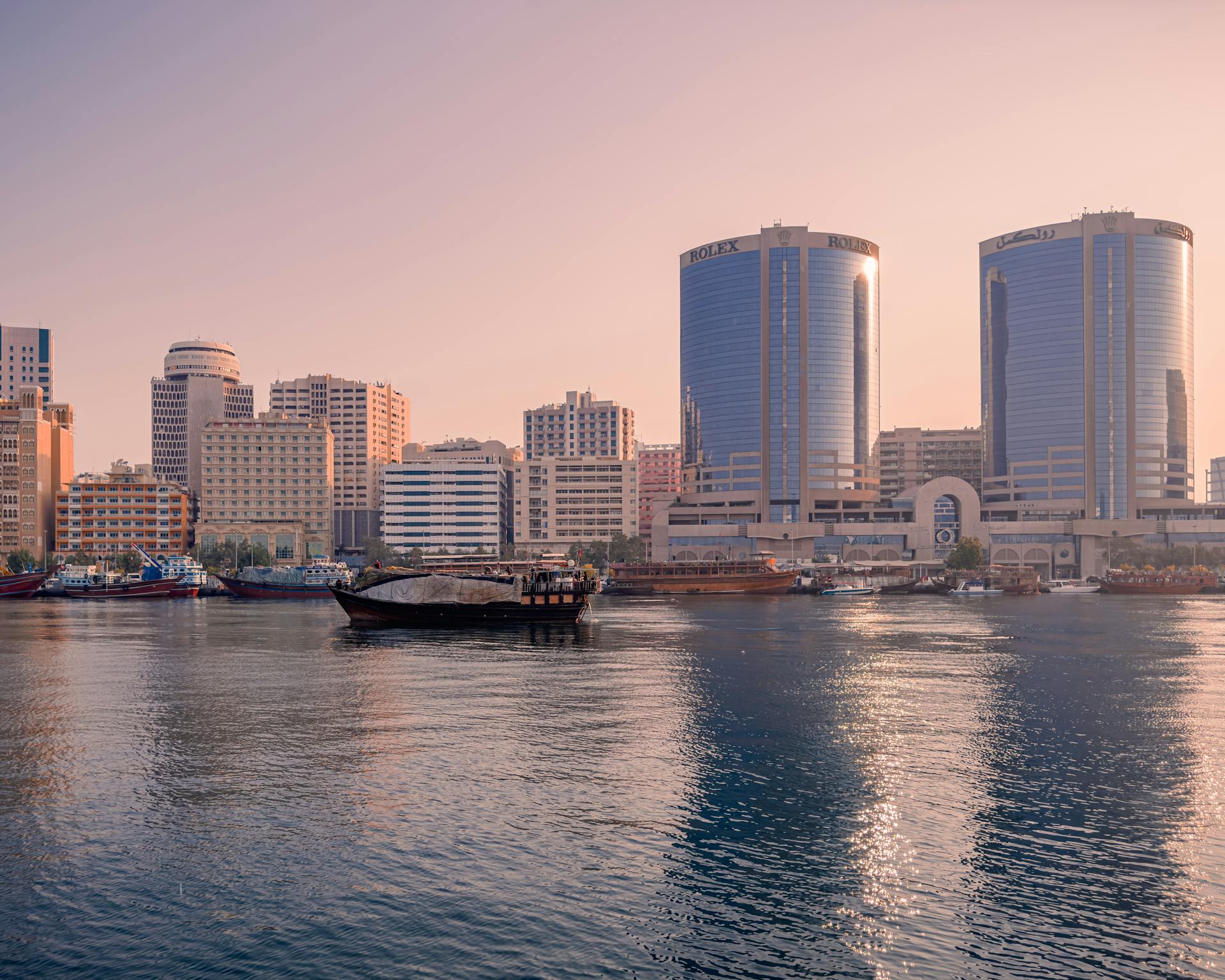 Modern buildings and traditional boats on Dubai Creek during a soft sunrise.