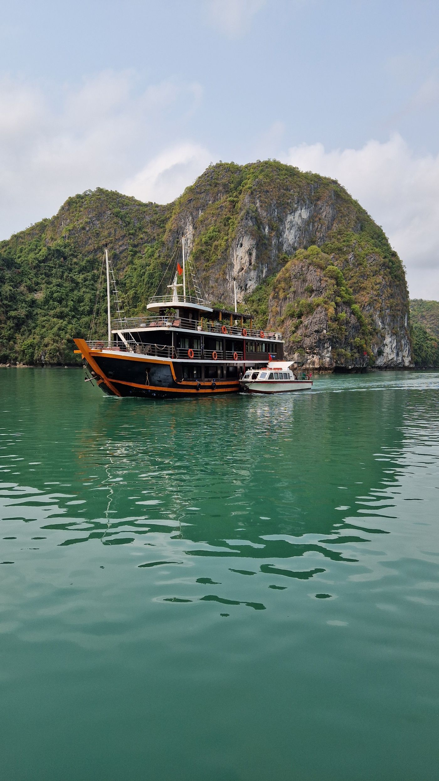 A two-deck wooden cruise junk sailing past a tall limestone island on green water in Ha Long Bay