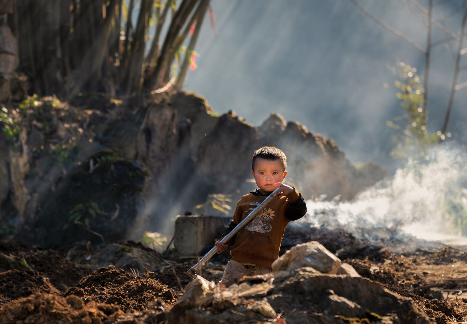 A young boy holding a tool in a sunny, rural, smoky outdoor setting.
