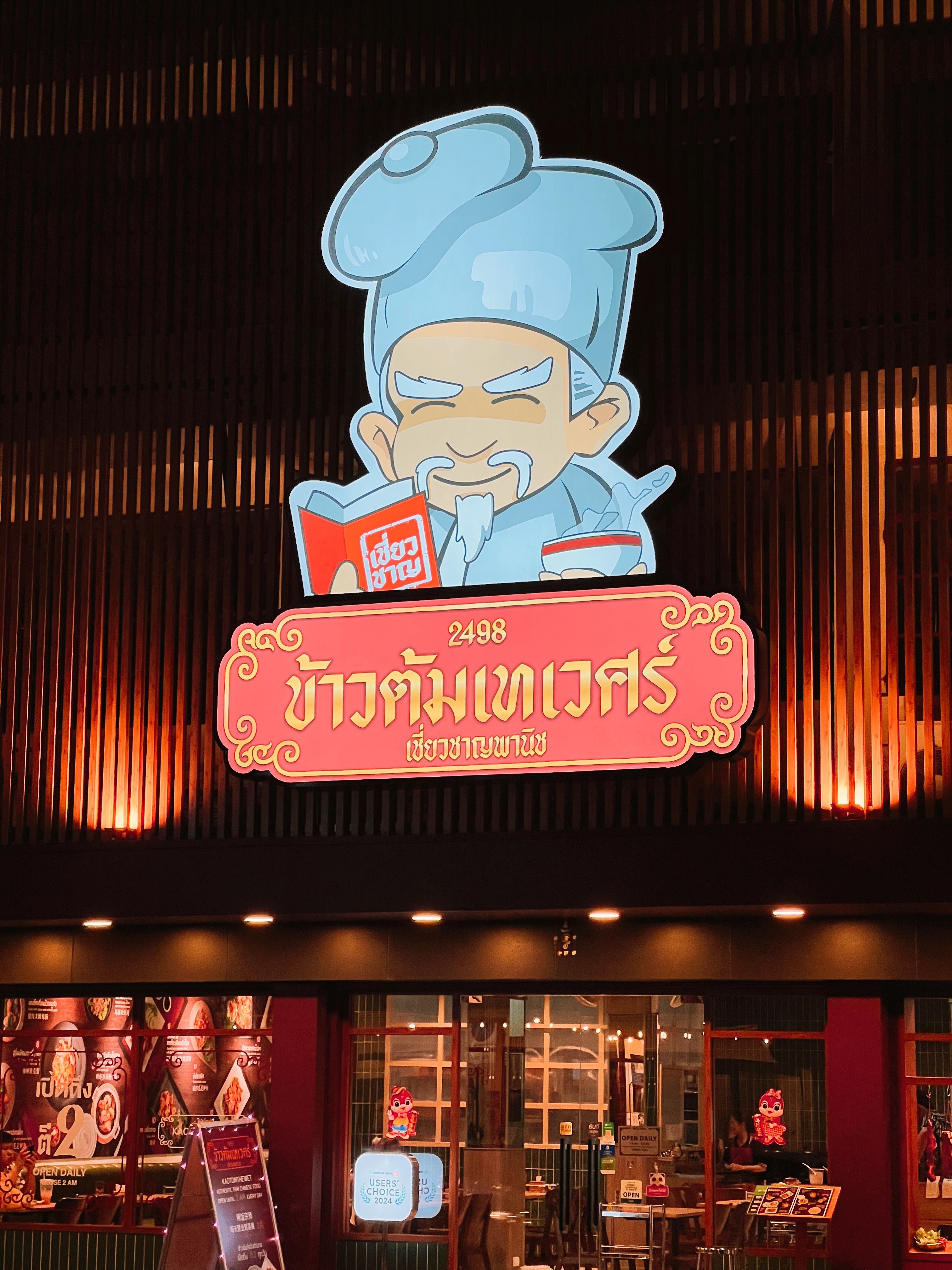 Vibrant Thai restaurant sign featuring cartoon chef above the entrance, warmly lit at night.