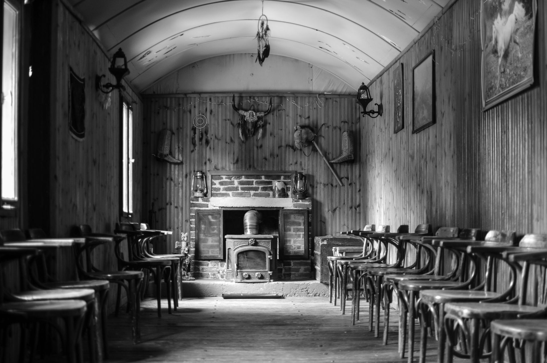 Black and white photo of a rustic cabin interior featuring a wood stove and vintage wooden chairs.