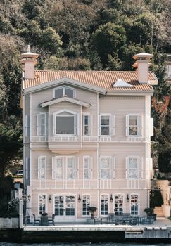 A beautiful three-story house with shutters by the waterfront, surrounded by lush greenery and trees.