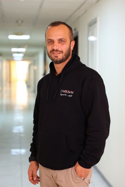 Smiling man in hoodie standing confidently in brightly lit hallway.