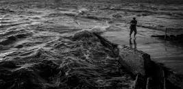 黑白图像中，一名渔民站在汹涌海浪的码头上