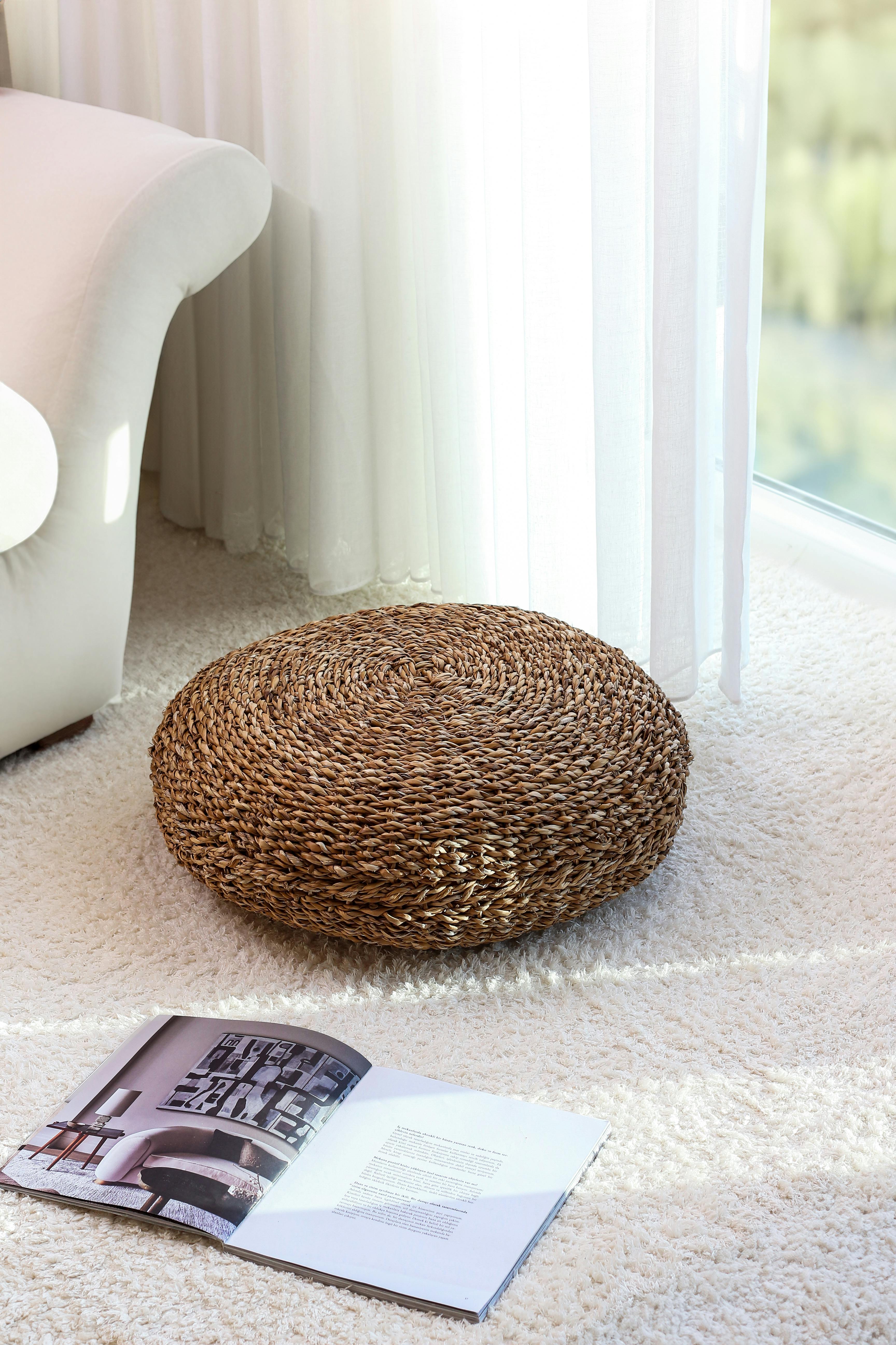 A stylish wicker pouf on shag carpet near a sofa and open magazine in bright living room.