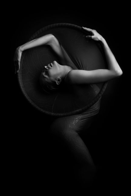 A striking monochrome photograph featuring a woman gracefully posing elegantly with a circular prop on a black background.