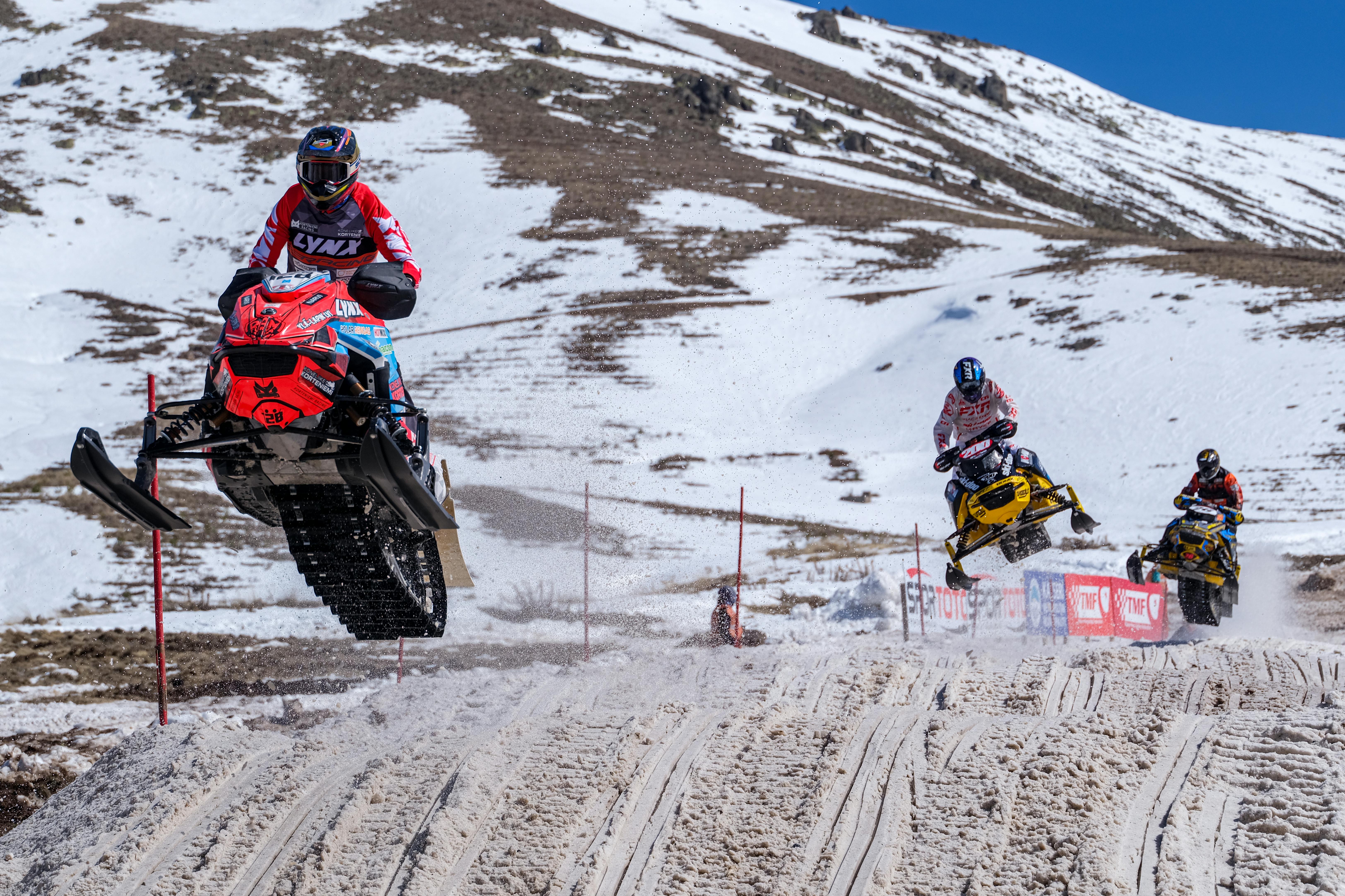 Exciting snowmobile racing in a snowy mountain setting with athletes in action, showcasing speed and skill.