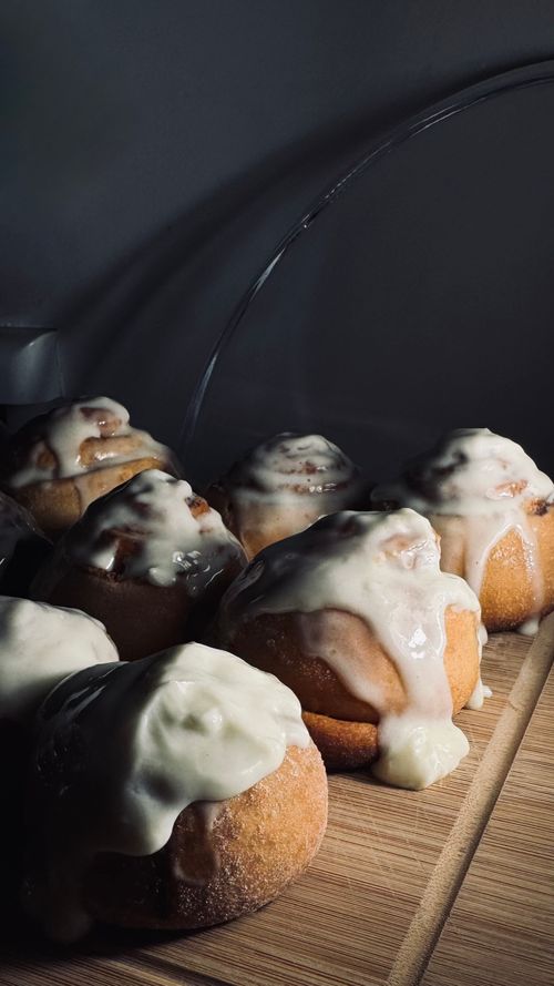 Free Close-up of warm, freshly baked cinnamon rolls covered in creamy white icing on a wooden board. Stock Photo
