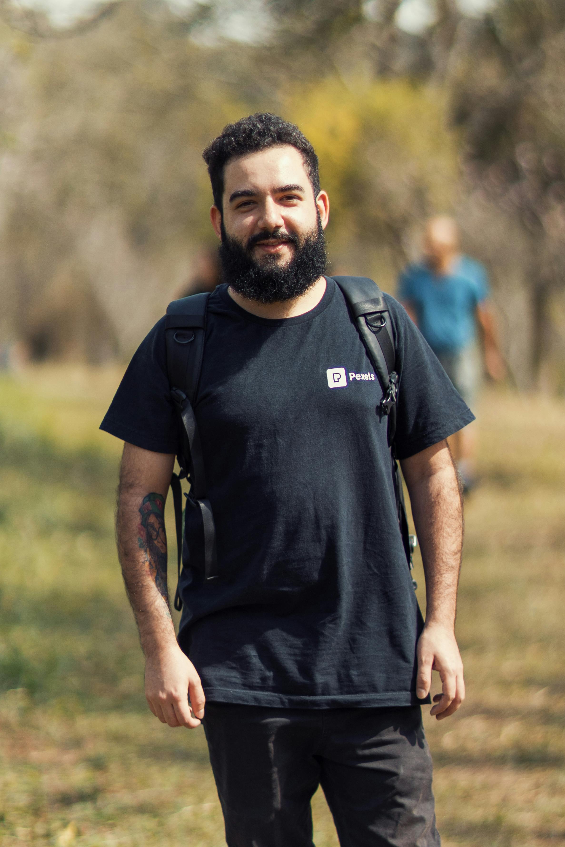 Bearded man smiling outdoors in casual black t-shirt with nature background, enjoying a sunny day.