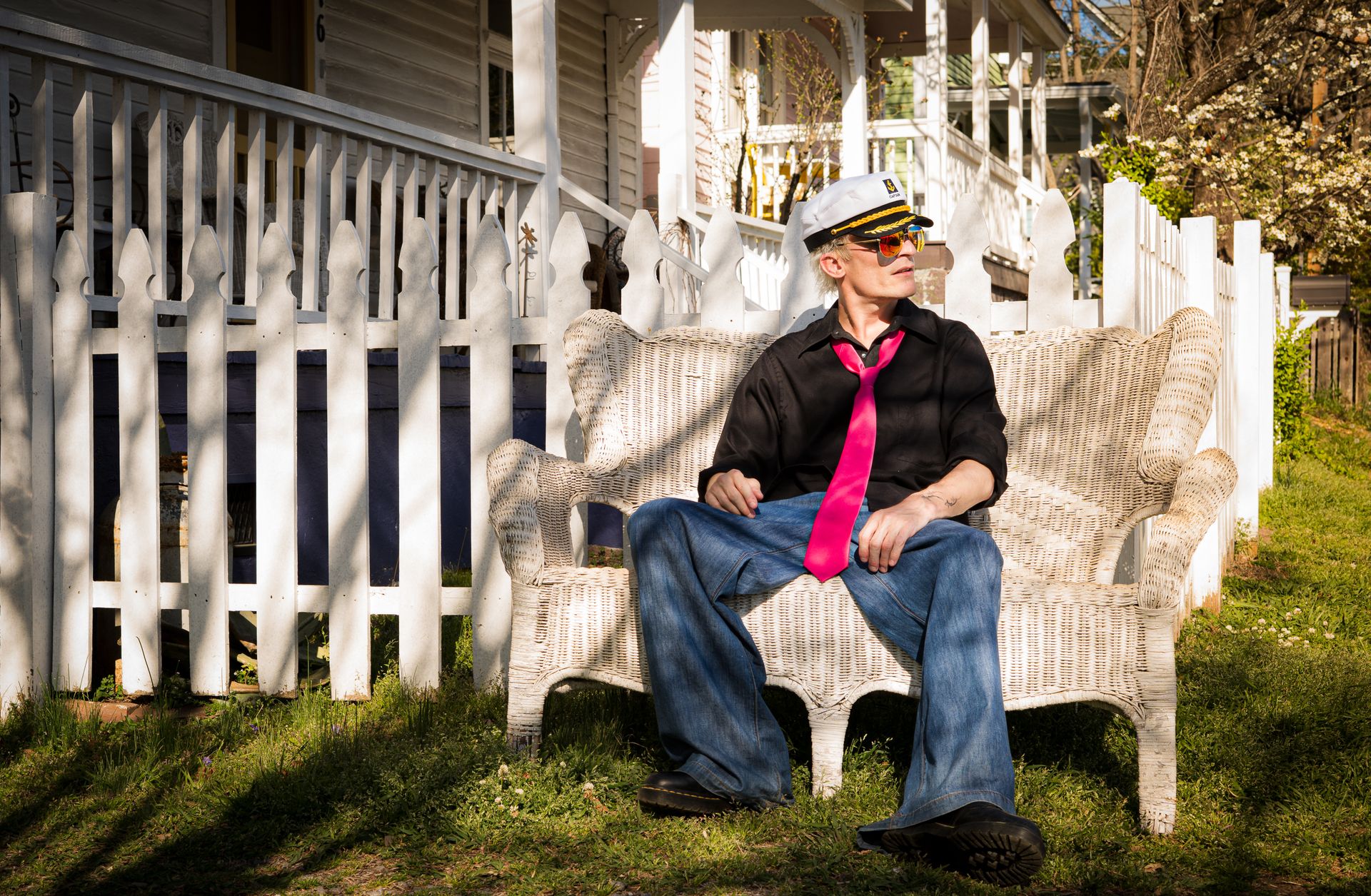 Man sitting on wicker bench in sailor hat outside house with white picket fence.