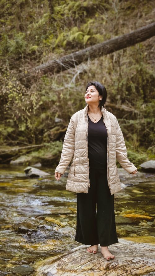 Free A woman in a warm jacket stands serenely by a rocky stream, appreciating nature. Stock Photo