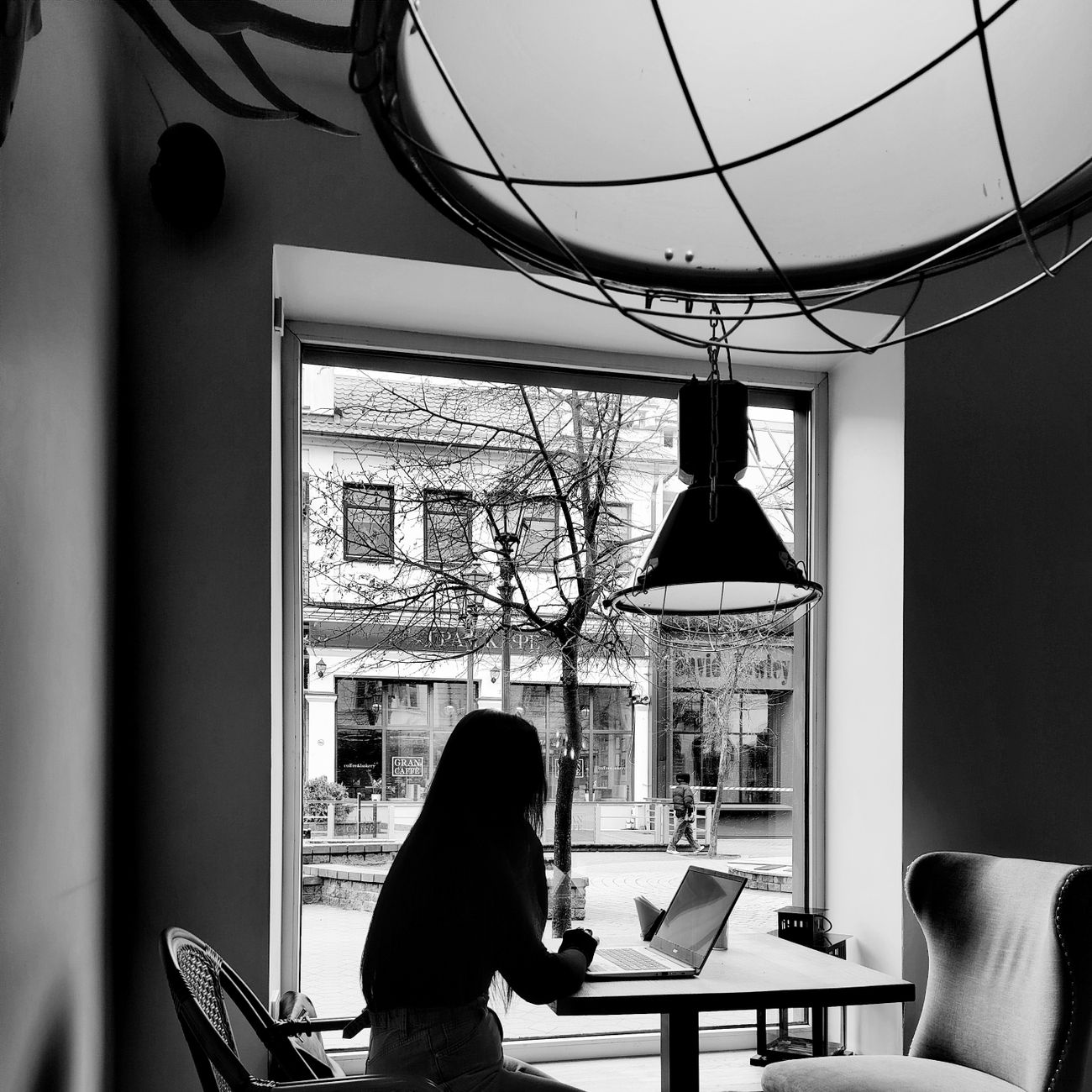 A woman using a laptop in a cozy cafe with a street view, captured in black and white.