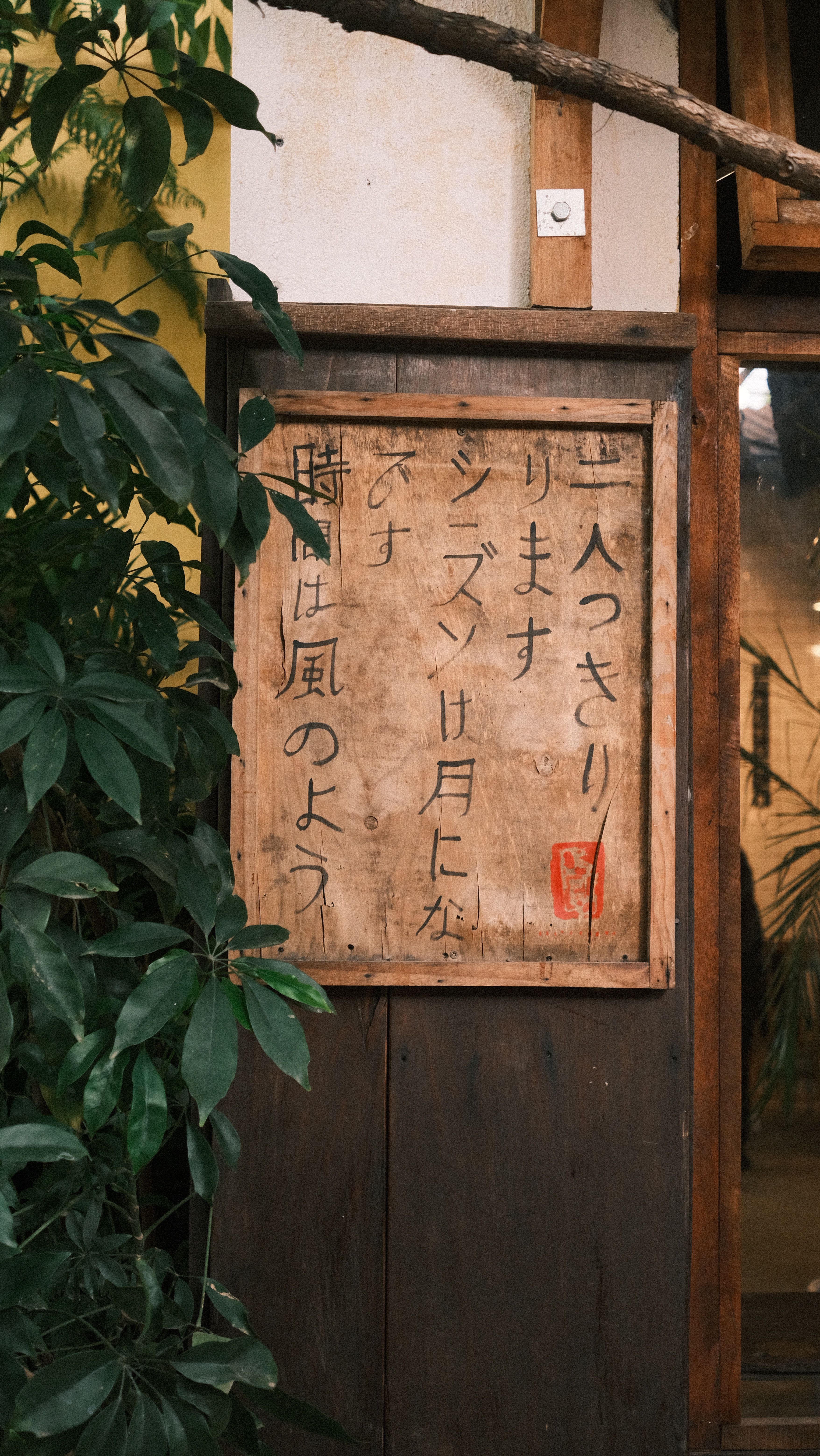 Japanese handwritten sign on a rustic wooden wall with lush green plants around.