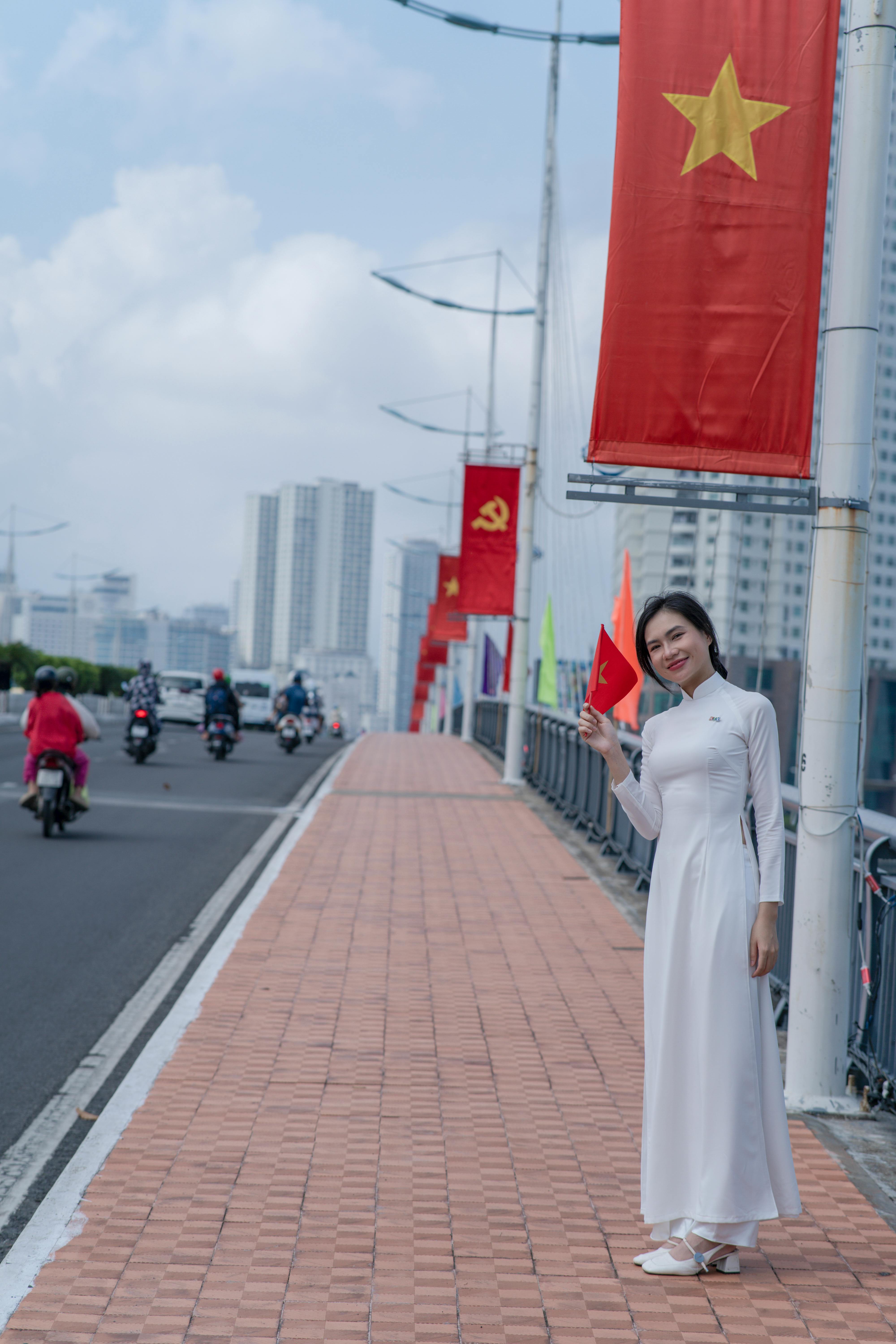 A woman in traditional dress holding a Vietnamese flag on a city bridge with flags lined up.