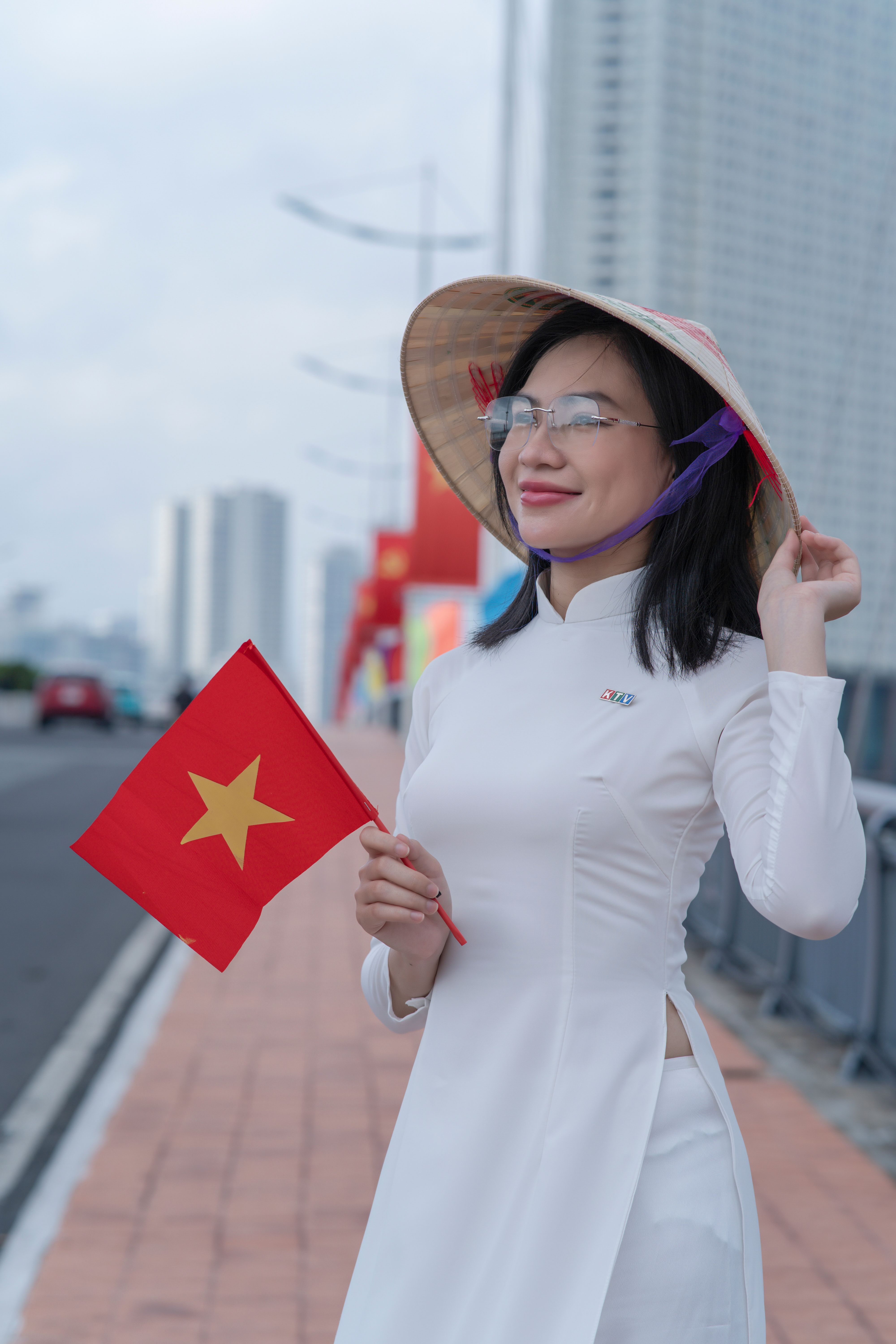 Smiling woman in white áo dài with Vietnamese flag in cityscape.