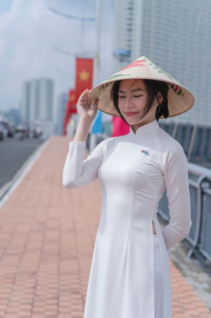 Woman in white Ao Dai and conical hat on urban sidewalk, serene.