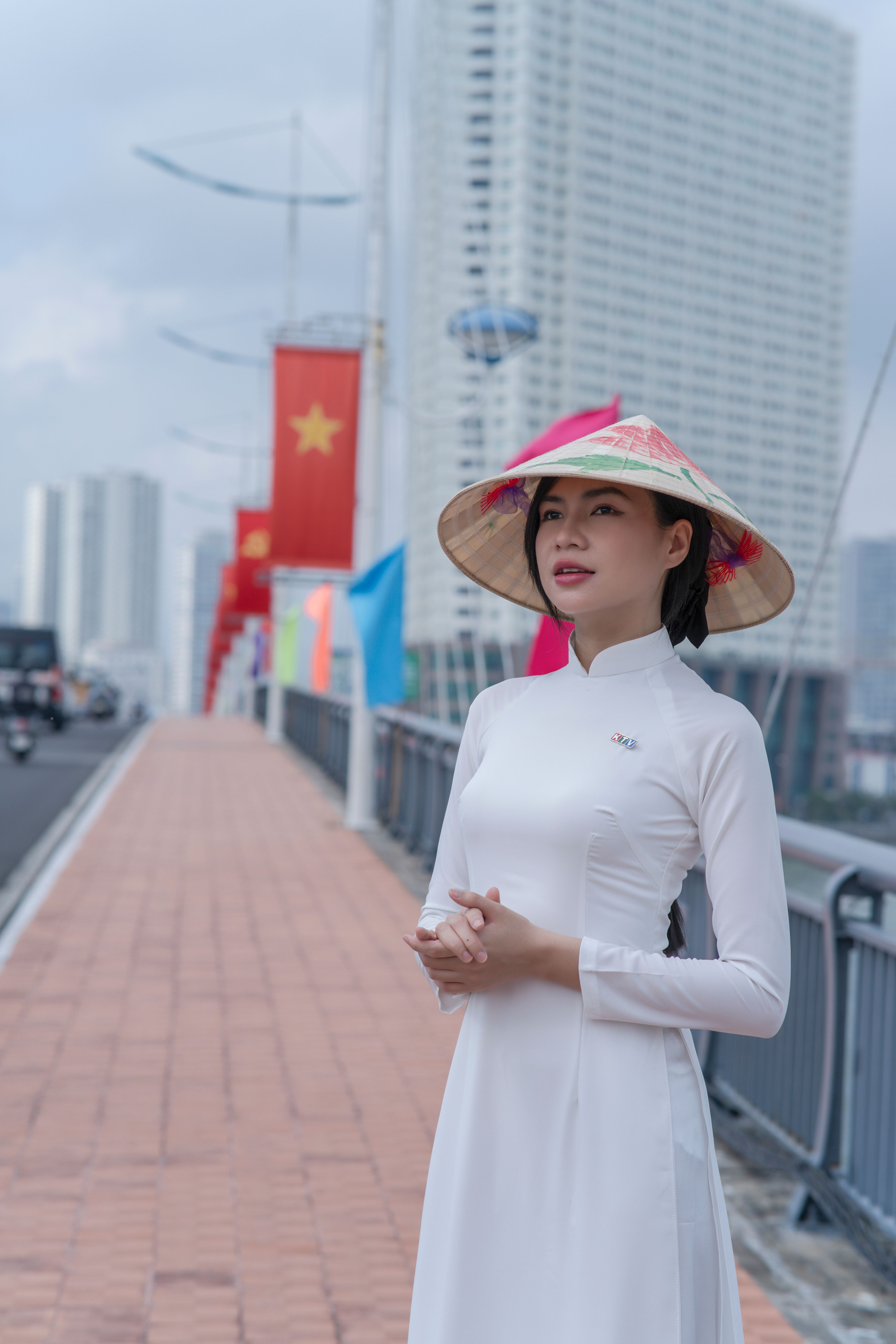 Woman in Ao Dai with Non La stands on city bridge with flags.
