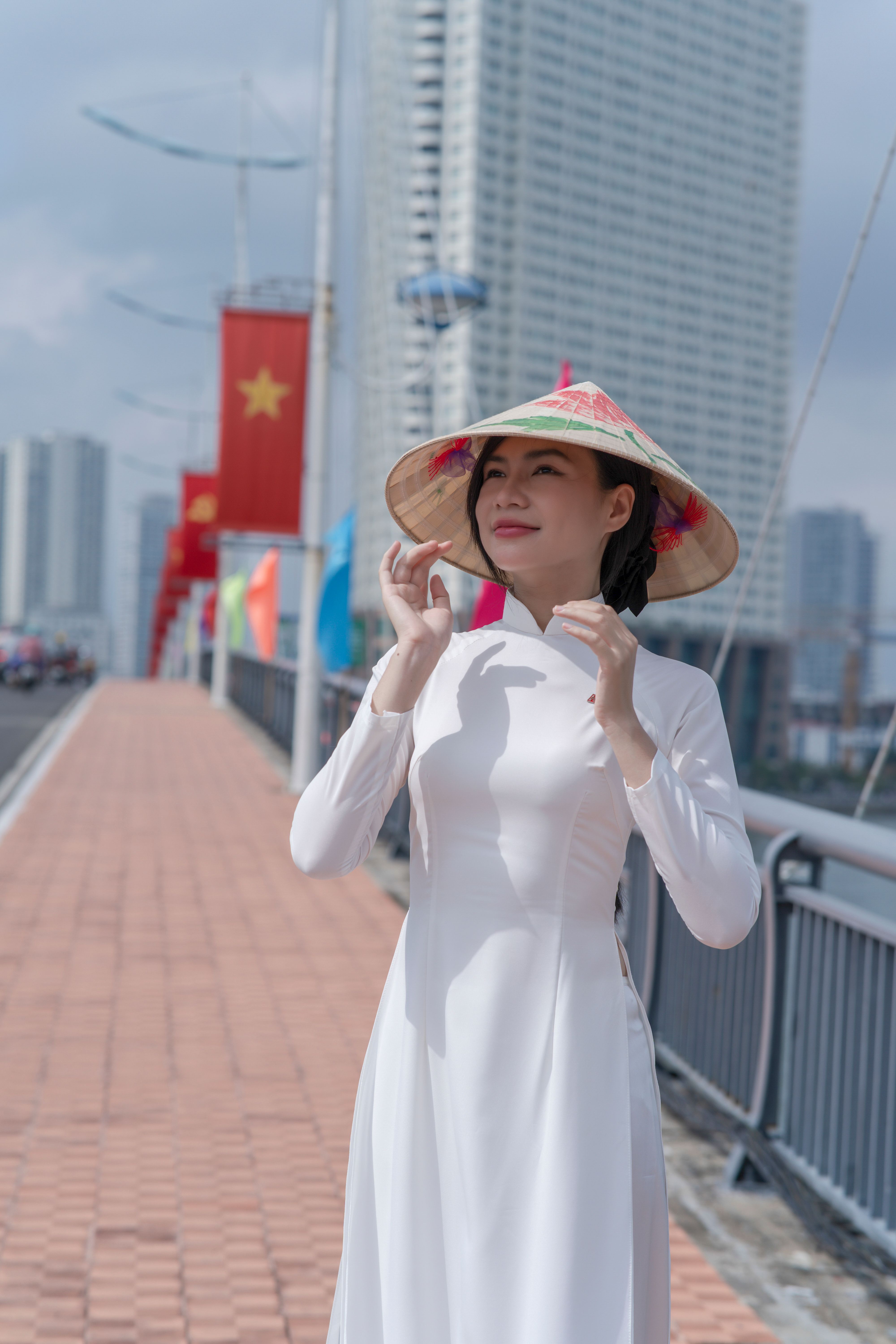 Elegant Vietnamese woman wearing ao dai and conical hat on a city bridge decorated with flags.