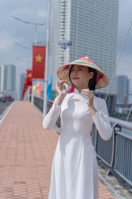 Elegant Vietnamese woman wearing ao dai and conical hat on a city bridge decorated with flags.