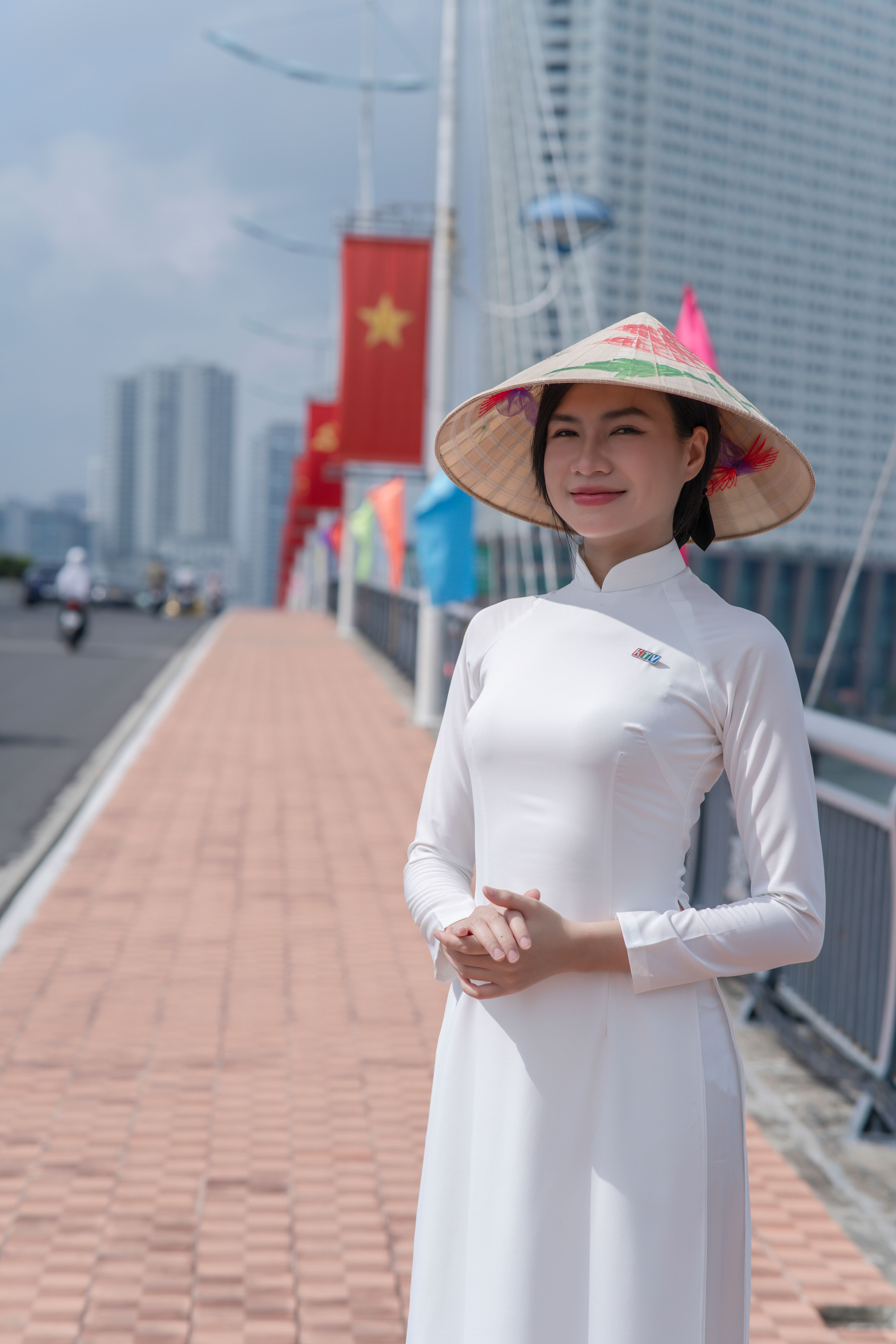 Woman in white Ao Dai with conical hat on bridge with flags in urban setting.