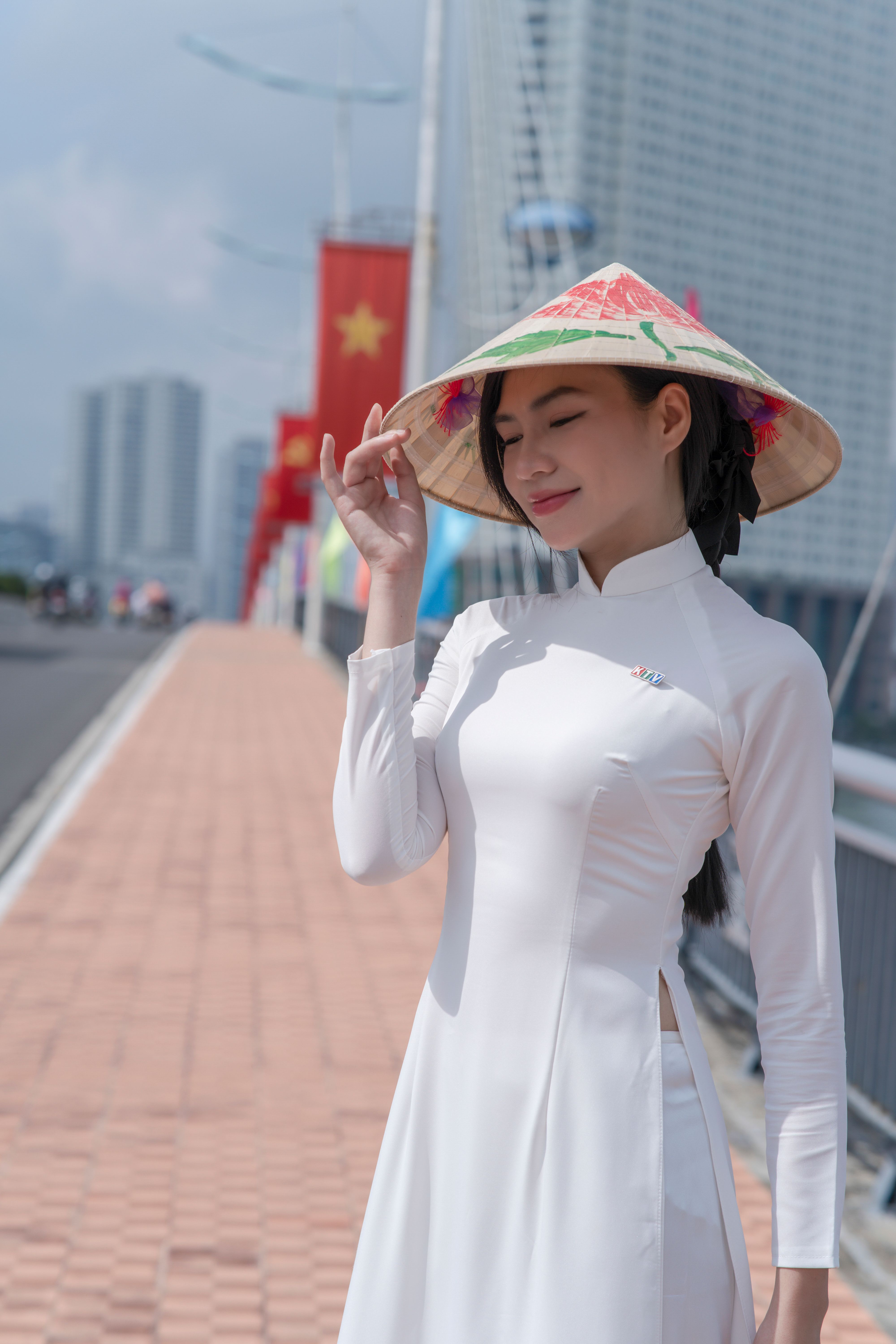 Smiling woman in white Ao Dai with hat on urban bridge under sunny sky.