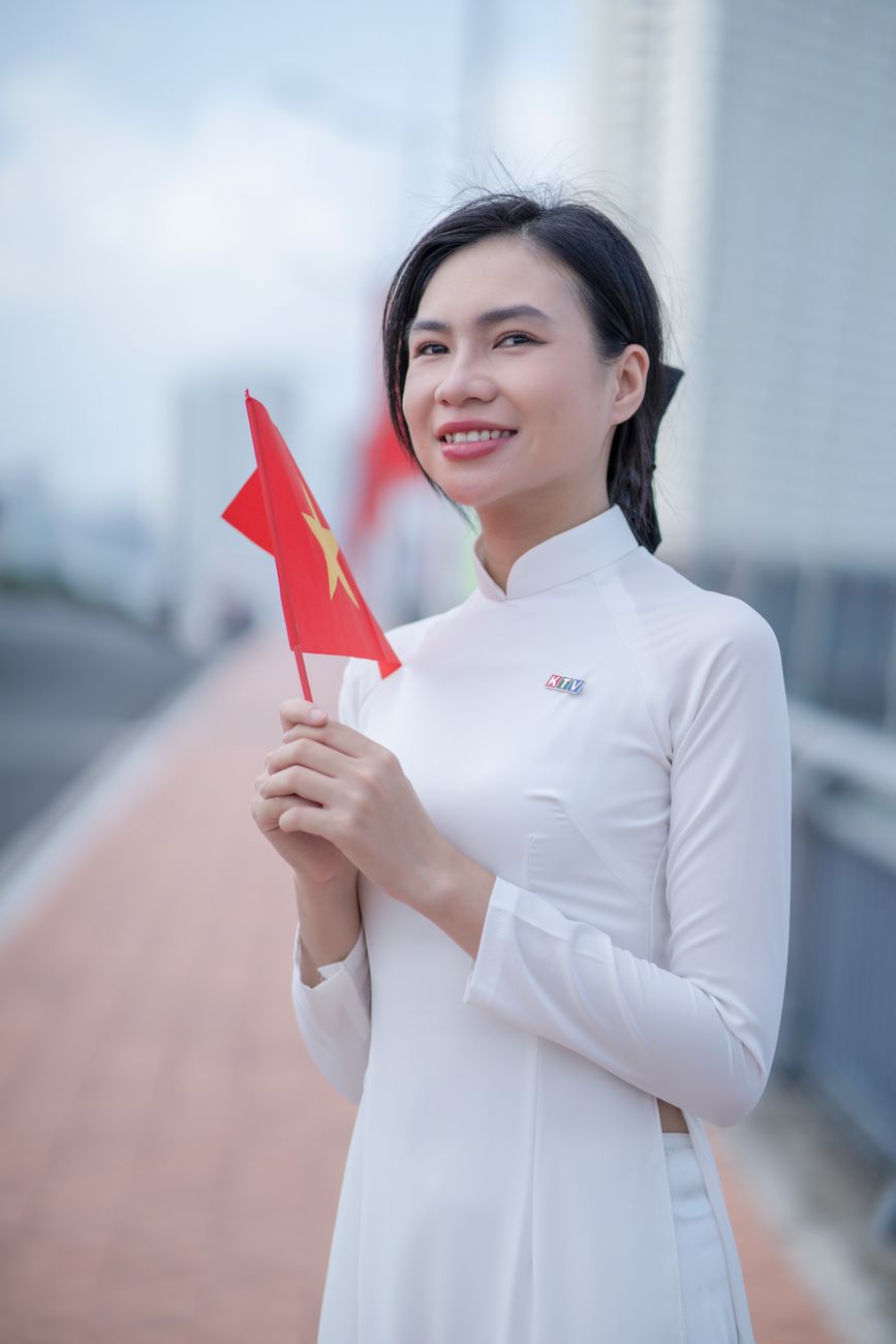 A smiling woman in traditional dress holds a Vietnamese flag, outdoors.