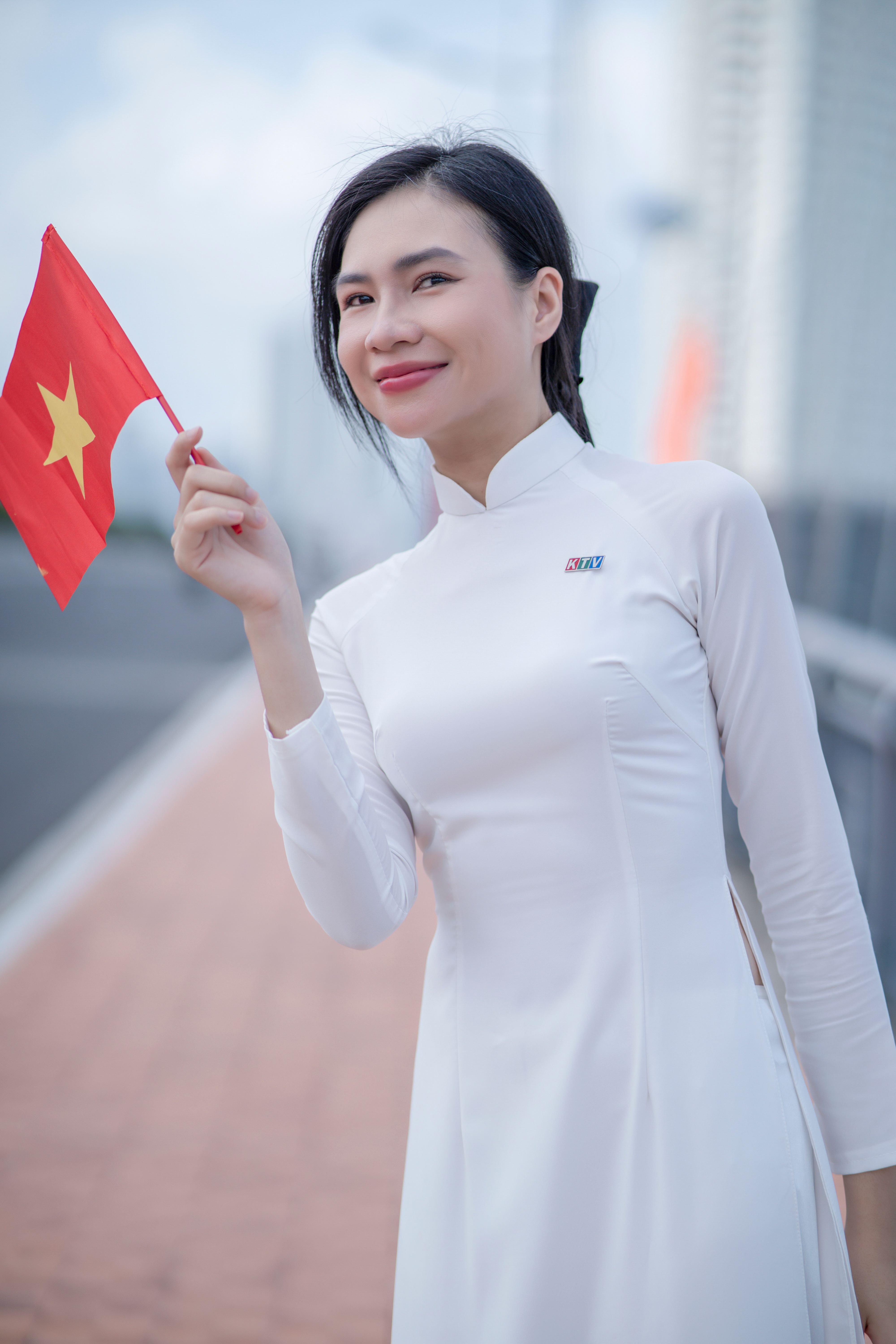 Smiling young woman in Ao Dai holding Vietnam flag outdoors.