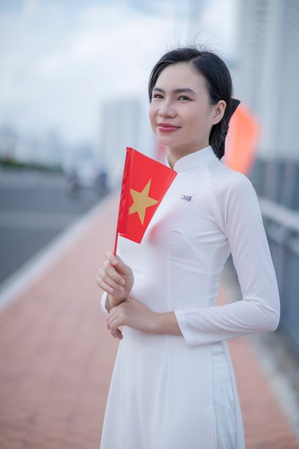 Smiling woman in traditional ao dai holding Vietnam flag outside.