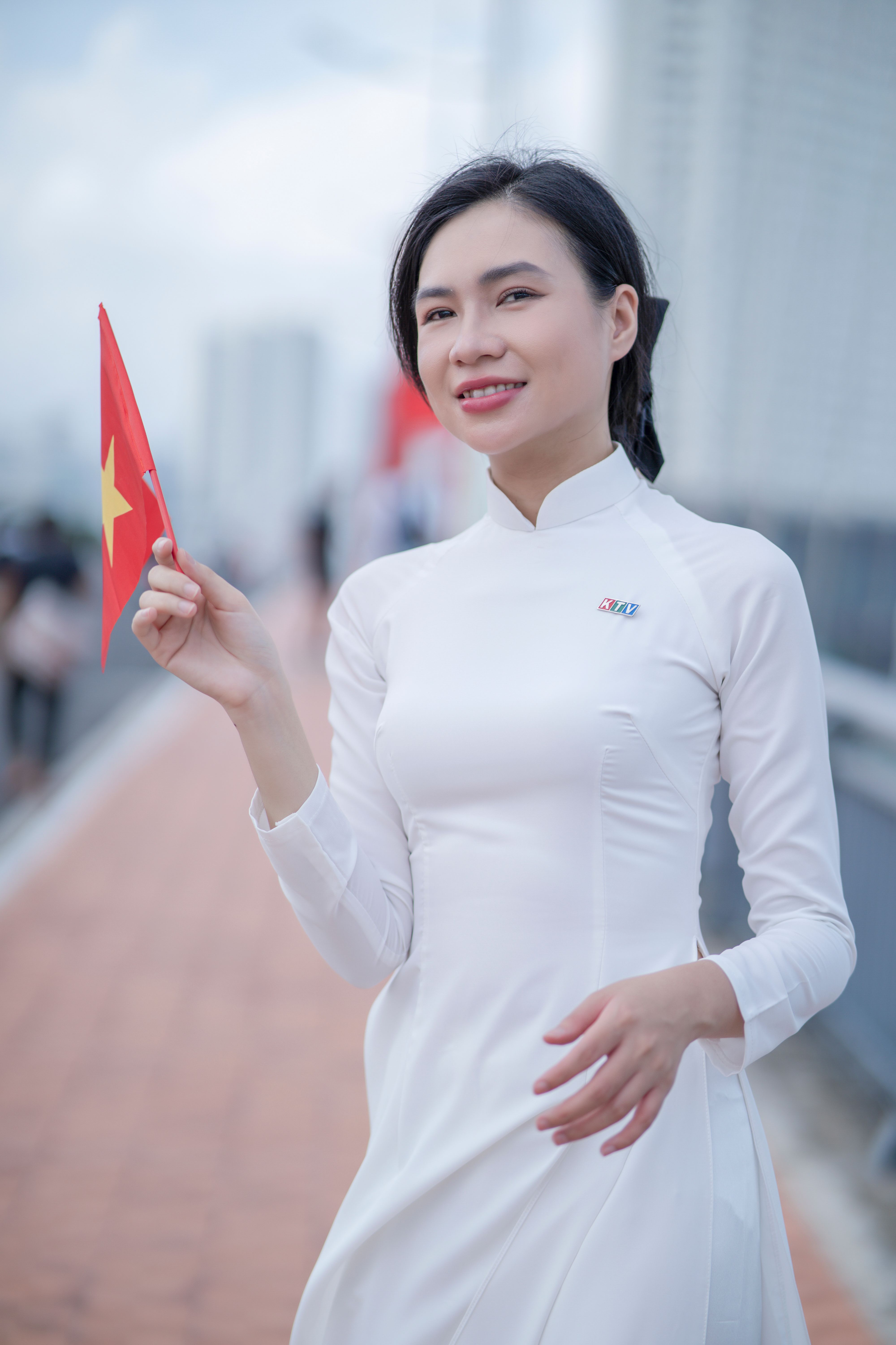 Woman in white áo dài holding a Vietnamese flag, smiling outdoors.