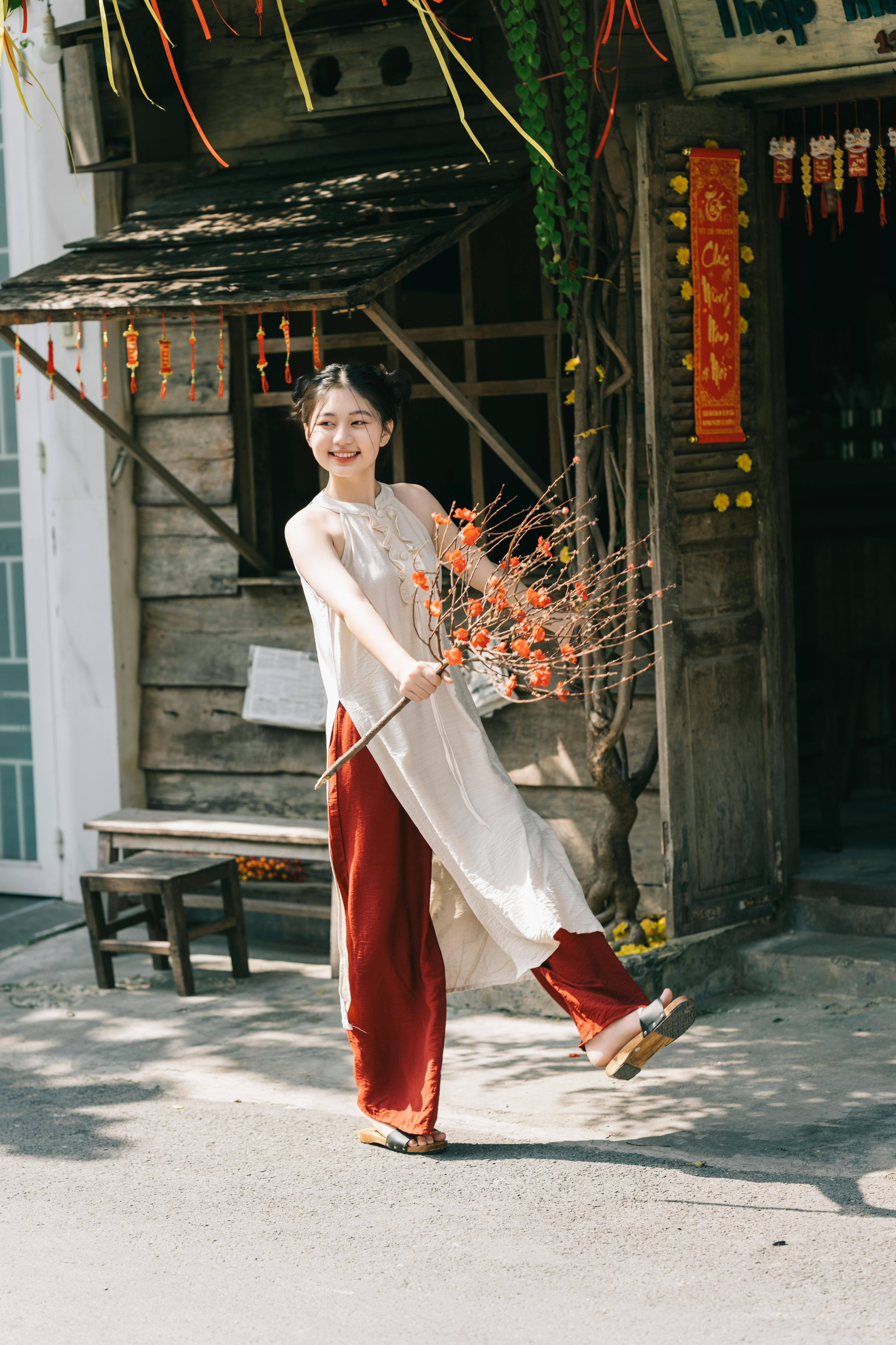 Joyful young woman in traditional attire celebrates Vietnamese New Year outdoors, holding vibrant flowers.