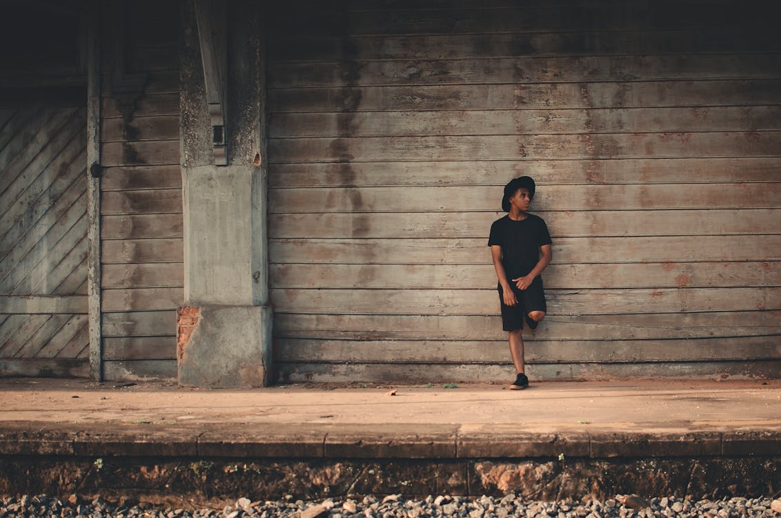 Photo of Man in Black T-shirt, Shorts, and Fedora Leaning on Wooden Wall Posing While Looking Away