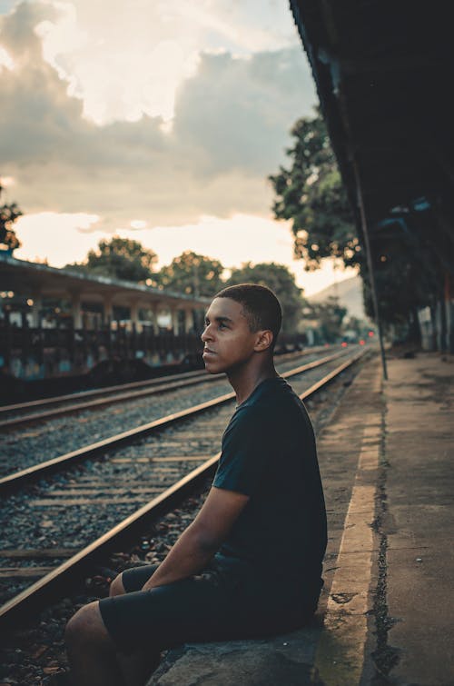 Photo of Man in Black T-shirt and Shorts Sitting on Train Platform Posing While Looking Away